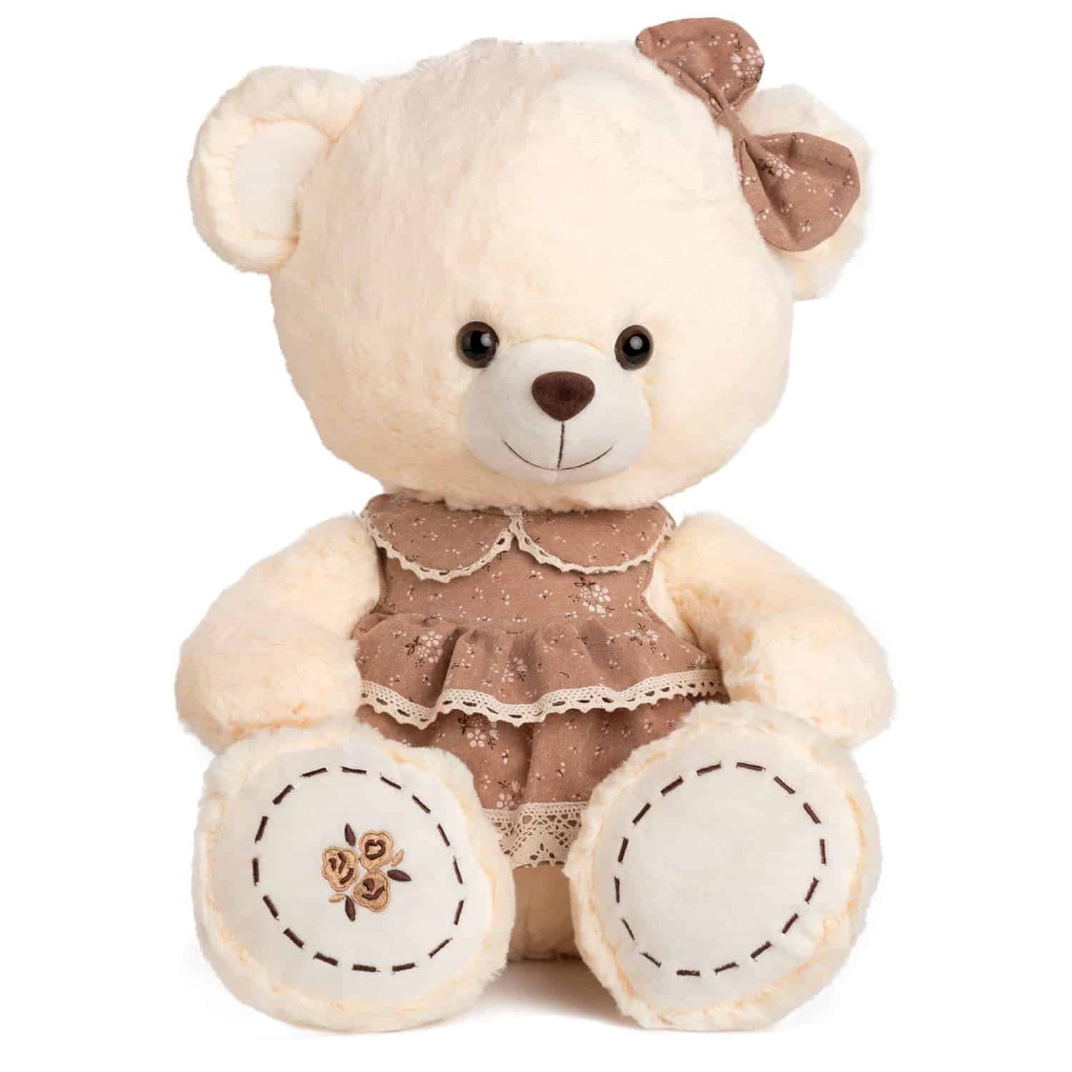 Меко плюшено мече с бежов цвят, облечено в сладко кафяво рокля и с цветна панделка на ухото, подходящо за подарък или декорация за детски празници.